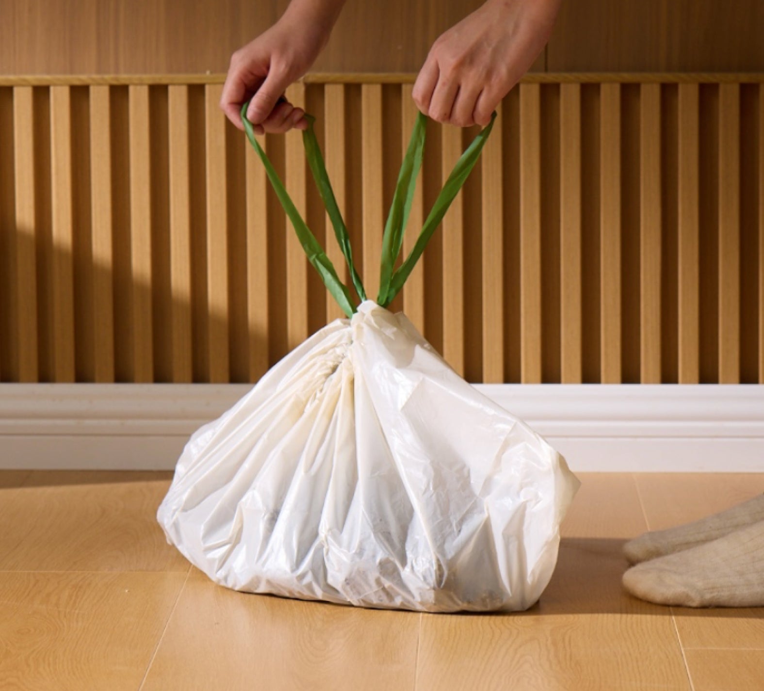 Petlibro white plastic trash bag with green drawstring handles being held by hands on wooden floor
