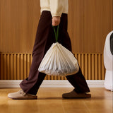 Petlibro lifestyle scene showing a person carrying a tied white trash bag with green handles indoors on wooden floor near a white pet product device