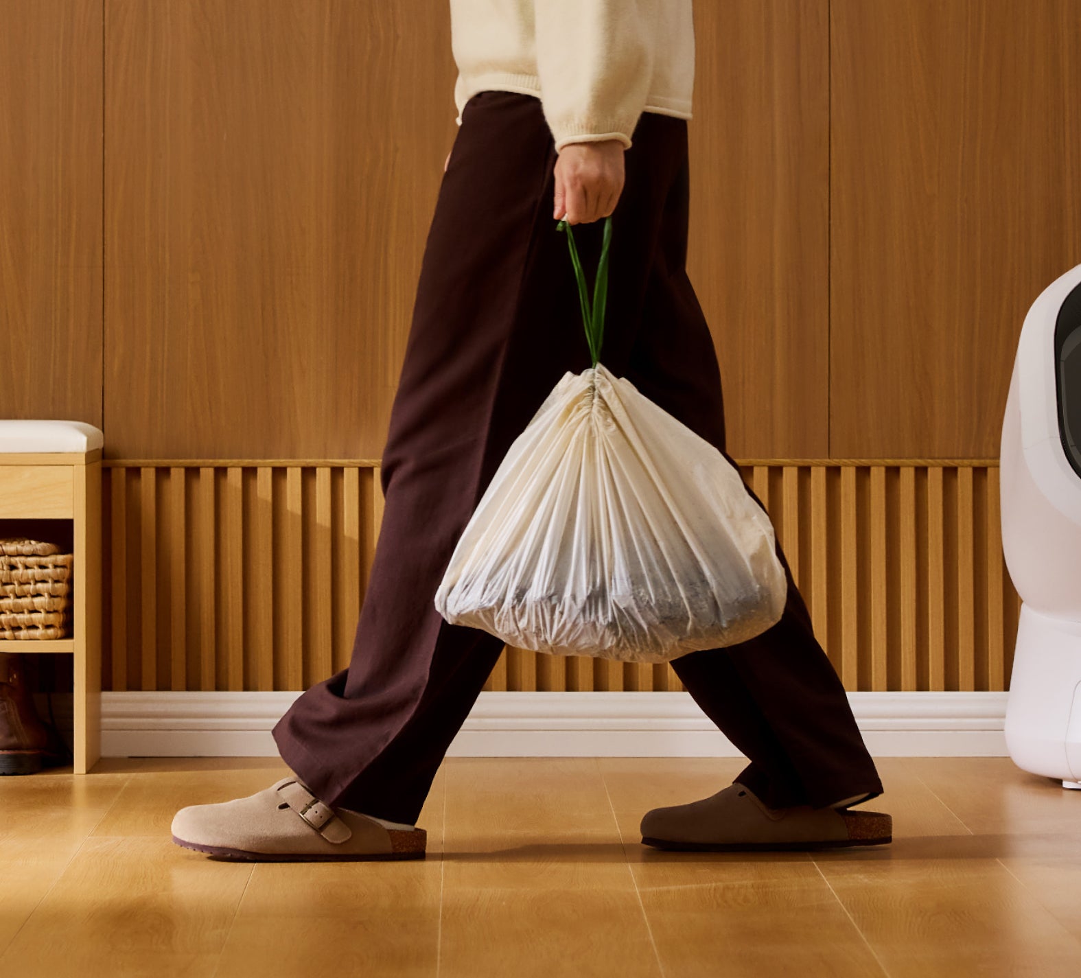 Petlibro lifestyle scene showing a person carrying a tied white trash bag with green handles indoors on wooden floor near a white pet product device