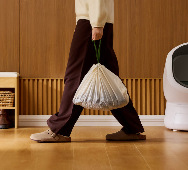 Petlibro lifestyle scene showing a person carrying a tied white waste bag near a white automatic cat litter box in a wooden interior room