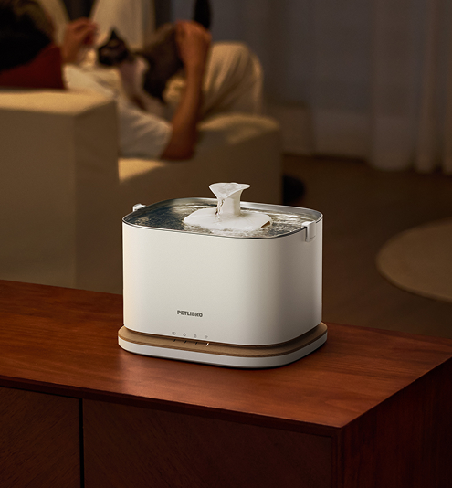 Petlibro PETLIBRO white pet water fountain on wooden table, flowing top spout in cozy living room scene, not a litter box
